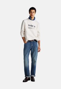 Sudadera blanca con texto gráfico en azul marino, jeans de mezclilla azules y zapatos de cuero marrón oscuro. Los jeans están enrollados en los puños, creando un aspecto informal.