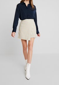 Blusa azul marino de botones y mangas largas, combinada con una minifalda a cuadros amarillos y blancos con dobladillo asimétrico, llevada con botas blancas hasta el tobillo.