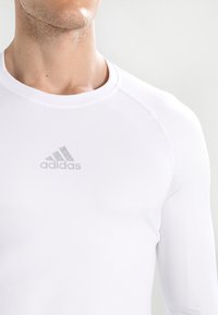 Balts garie piedurknēm sporta krekls, kas izgatavots no elastīga materiāla, ar apaļu izgriezumu un diskrētu pel�ēku Adidas logotipu uz krūtīm.