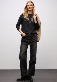 Sort sweatshirt med teksten "PARIS CLUB DE LA RUE", kombineret med højtaljede mørke jeans. Modellen har sorte ankelstøvler på. Blød stoftekstur.