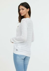 Pull en tricot blanc avec motif en zigzag, léger, avec des poignets et un ourlet côtelés, porté avec un jean bleu. Étiquette de logo sur le côté visible.