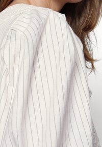 Gros plan d'une personne portant un vêtement blanc avec de fines rayures verticales grises et des cheveux bruns visibles sur un fond clair.