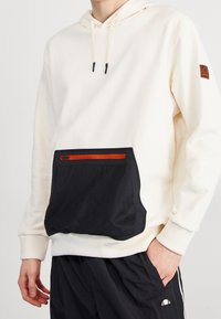 Sudadera con capucha de tela crema que presenta un gran bolsillo de malla negra con un zipper rojo. Incluye puños y dobladillo de canalé.