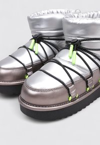Bottes argentées et gris métalliques avec un revers en peluche, dotées de lacets noirs et d'accents verts. Semelle épaisse en caoutchouc noir avec des coutures distinctes.