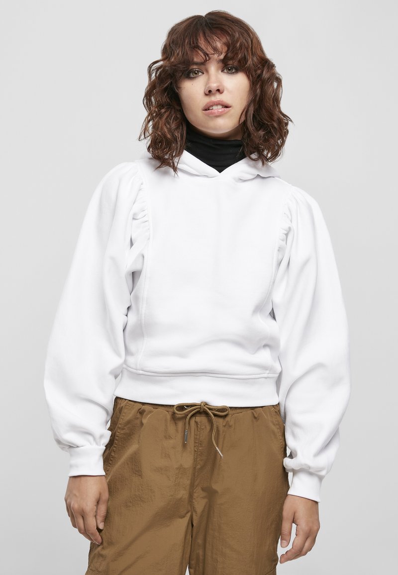 Jeune femme aux cheveux bruns bouclés portant un sweat à capuche blanc à manches bouffantes sur un haut noir et un pantalon marron à cordon, debout devant un fond uni.