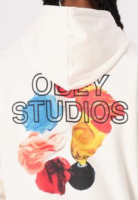 Cream hoodie featuring a large graphic on the back with colorful paint swirls and bold black text reading "OBEY STUDIOS."
