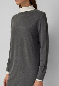 Eine Frau trägt ein graues langärmliges Strickkleid über einem weißen Oberteil mit hohem Kragen und steht vor einem einfarbigen Hintergrund.