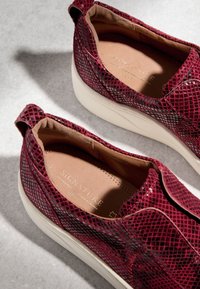 Paire de baskets à enfiler bordeaux à motif cuir de serpent avec semelles blanches et semelles intérieures beige rembourrées sur surface grise.