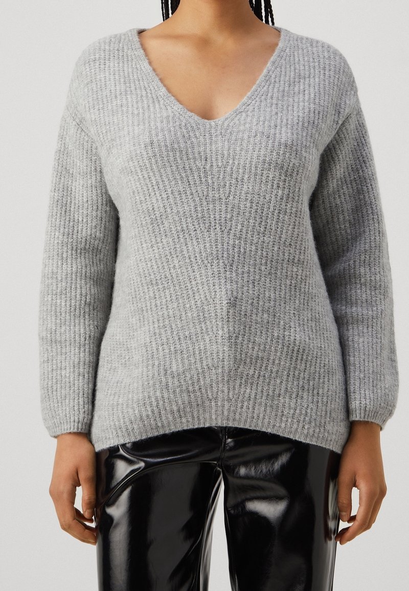 Szary sweter dziany z dekoltem w kształcie litery V, długimi rękawami i prążkowaną fakturą; zestawiony z błyszczącymi czarnymi spodniami.