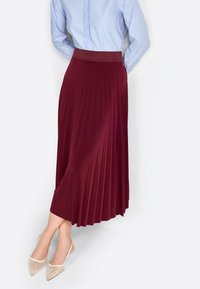 Falda plisada de longitud media en tela burdeos, con una cintura elástica texturizada y un drapeado suave y fluido. Combinada con zapatos de talón destalonado en color beige.