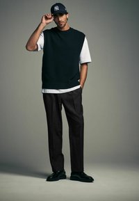 Dark green knitted sweater vest over a white t-shirt, paired with black pinstriped trousers and black lace-up shoes. Navy baseball cap worn.