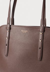 Bruine leren tote bag met textuur, dubbele handvatten, gouden logo details en uitgesproken stiksels. Heeft een gestructureerde, rechthoekige vorm.