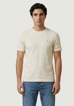 Hombre de pie mirando al frente, vistiendo una camisa de manga corta color crema, jeans azules y zapatillas blancas, sobre un fondo gris liso.