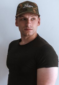 Kamuflázs baseball sapka "Blackskies" logóval; fekete, testhezálló póló. Rövid haj, egy világos falnak támaszkodik. Fókuszban a fej és a vállak.