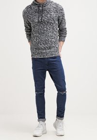 Grau-schwarzer gestrickter Kapuzenpullover mit strukturiertem Muster, kombiniert mit eng anliegenden blauen Jeans mit Used-Details und weißen Turnschuhen.