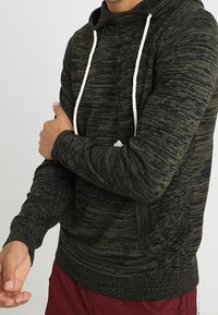 Uomo in felpa nera e grigia a motivo con lacci bianchi che aggiusta il maniche sinistro, indossando pantaloni sportivi bordeaux.