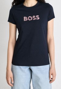 Žena v černém tričku se nápisem "BOSS" poskládaným z růžových a bílých květinových flitrů, v kombinaci se světle modrými džíny.