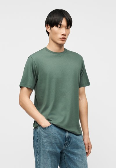 Νεαρός άνδρας με μαύρα μαλλιά που φοράει ένα απλό πράσινο κοντομάνικο μπλουζάκι και μπλε τζιν, στέκεται με το ένα χέρι στην τσέπη.