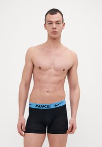 Sorte Nike boxershorts med en lys blå midjebånd som har merkevarenavnet. Glatt tekstur og tettsittende design. Modellen er vendt mot kameraet.