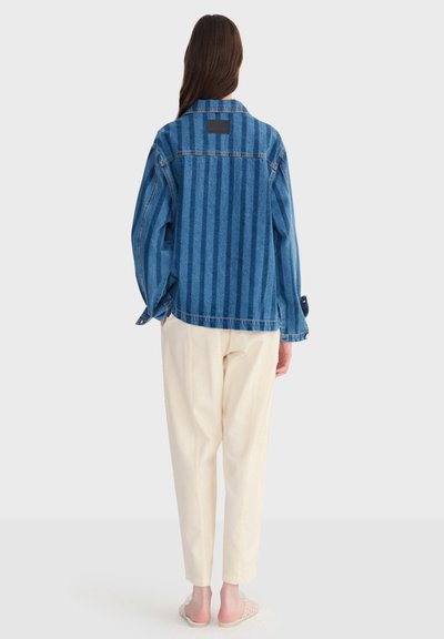 Femme aux longs cheveux bruns portant une veste en jean rayée bleue, un pantalon crème et des chaussures transparentes sans lacets, debout de dos.