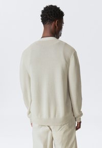 Pull en tricot couleur crème avec une coupe décontractée, des poignets côtelés et une texture lisse, présenté de dos sur un fond uni.