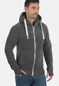 Gray fleece zip-up hoodie with white drawstrings, side pockets, and a red logo on the chest. Soft texture, casual fit.