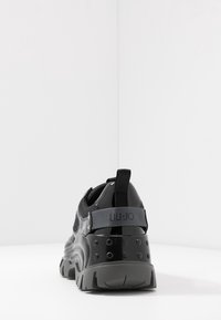 Černá sportovní bota s texturovanou síťovinou, lesklými akcenty a robustní podešví. Na zadní straně se nachází šedý pásek s logem "LIU JO".