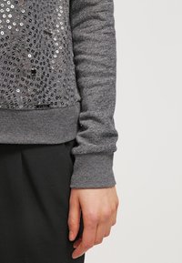 Bras et main féminins à côté d'une chemise grise à manches longues avec des ornements en sequins et un pantalon noir sur un fond clair uni.