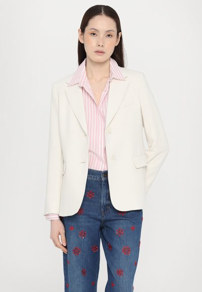 Blazer bianco su misura con colletto a revers, abbinato a una camicia a righe rosa e jeans azzurri arricchiti da motivi floreali rossi.
