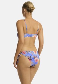 Frau mit einem Dutt, die einen blauen Blumenbikini trägt, von hinten gesehen. Der Bikini hat lebendige Blumen und verstellbare Bänder an den Seiten.