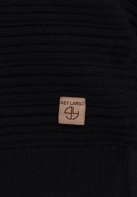 Tissu noir texturé avec des nervures horizontales, comportant une étiquette en cuir beige portant l'inscription "KEY LARGO" et un logo circulaire.