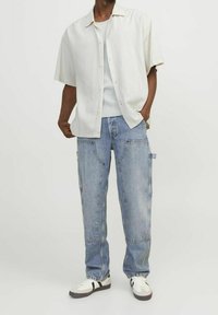 Lichtbeige overhemd met korte mouwen en knopen, gedragen over een witte geribbelde tanktop, gecombineerd met lichtblauwe wijde jeans en witte sneakers met zwarte accenten.