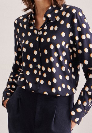 Vrouw draagt een donkerblauw overhemd met witte en oranje polkadotjes, gestopt in een donkerblauwe broek, handen in de zakken.