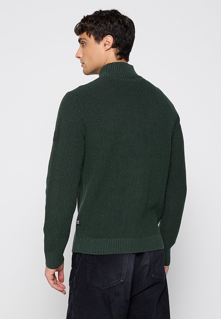 Zielony sweter z dzianiny o wysokim kołnierzu, z fakturowanym wzorem, ściągaczem u dołu i łatami na łokciach, pokazany od tyłu na modelu.