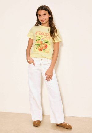 Fată tânără cu păr lung castaniu, purtând un tricou galben cu grafică de citrice, pantaloni albi cu croială largă și pantofi maro slip-on, stând în picioare lângă un perete bej.