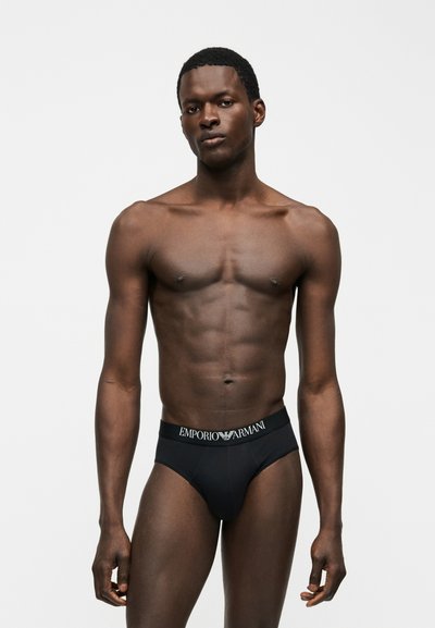 Αθλητικός σκουρόχρωμος άνδρας στέκεται φορώντας μαύρα μποξεράκια Emporio Armani σε ένα απλό ανοιχτό γκρι φόντο.