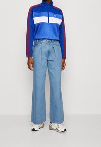 Chaqueta azul con cremallera, rayas blancas y rojas, combinada con jeans azul de corte holgado y zapatillas multicolores sobre un fondo gris.