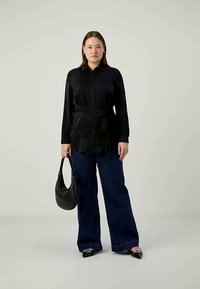 Melna blūze ar jostu, izgatavota no gluda auduma, kombinēta ar brīvām tumši zilām džinsām un melnu plecu somu. Apaviem ir norādīta pirksta forma.