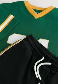 Groene mesh jersey met oranje en witte accenten, voorzien van een wit nummer, gecombineerd met zwarte sweatpants met witte trekkoord en afwerking.