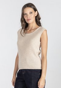 Beżowy bezrękawnik z okrągłym dekoltem, wykonany z miękkiego materiału. Prążkowany dół w talii, zestawiony z ciemnoniebieskimi dżinsami.