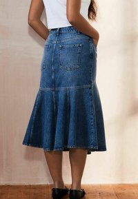 Denim midi nederdel med en bredere kant, rå kant og to baglommer. Farven er mellem blå med en subtil tekstur. Sorte sko og trægulv synligt.