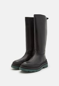 Bottes montantes en cuir noir avec une finition texturée, bout rond et une semelle en caoutchouc épaisse de couleur verte. Elles présentent des détails de couture et une languette pour faciliter l'enfilage.