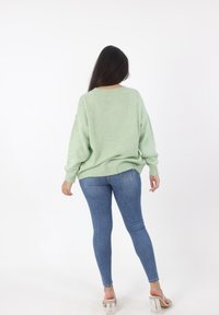 Lichtgroene oversized trui met geribde manchetten en zoom, gedragen over strakke blauwe jeans. De achterkant toont een relaxte pasvorm en zachte textuur.