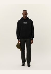 Felpa nera con cappuccio e tasca frontale, caratterizzata da un logo testuale bianco. Abbinata a pantaloni in denim scuro e un cappellino verde tenuto in una mano.