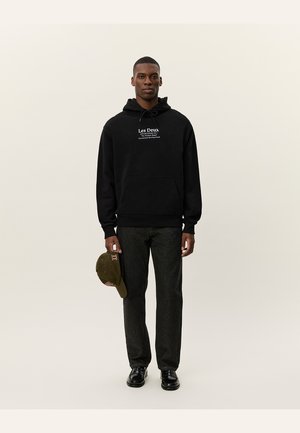 Svart hettegenser med frontlomme, som har en hvit tekstlogo. Kombinert med mørk denimbukser og en grønn caps i den ene hånden.