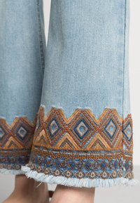 Vaaleansiniset farkkulegginssit, joissa on monimutkaista geometristä kirjontaa oranssilla, ruskealla ja sinisellä lahkeissa, ja hapsuviimeistely.