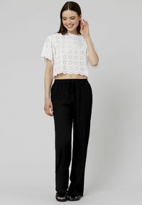 Top court blanc avec un motif de broderie florale, bords festonnés, associé à un pantalon noir ample et des sandales noires.