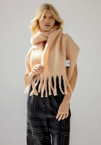 Châle en mélange de laine pêche avec des bords frangés, texture douce ; porté sur une tenue noire avec des motifs géométriques blancs. Étiquette visible sur le châle.