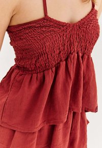 Žena v bezramienkových šatách v tehlovooranžovej farbe so zberaným telom a vrstvenou sukňou, zobrazená od ramien po stred stehna.