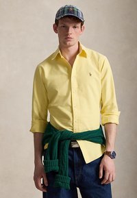 Homme portant une chemise jaune à boutons avec manches retroussées, jean foncé, pull vert noué à la taille, casquette à carreaux et montre-bracelet.